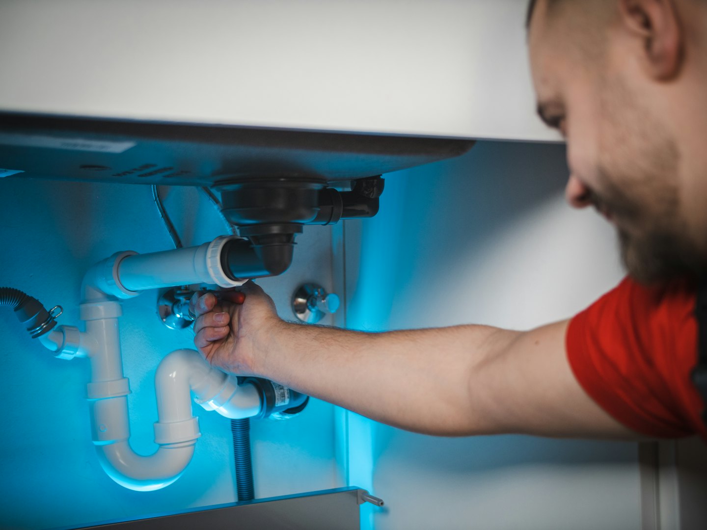Man in red shirt works on pipes under a sink.
